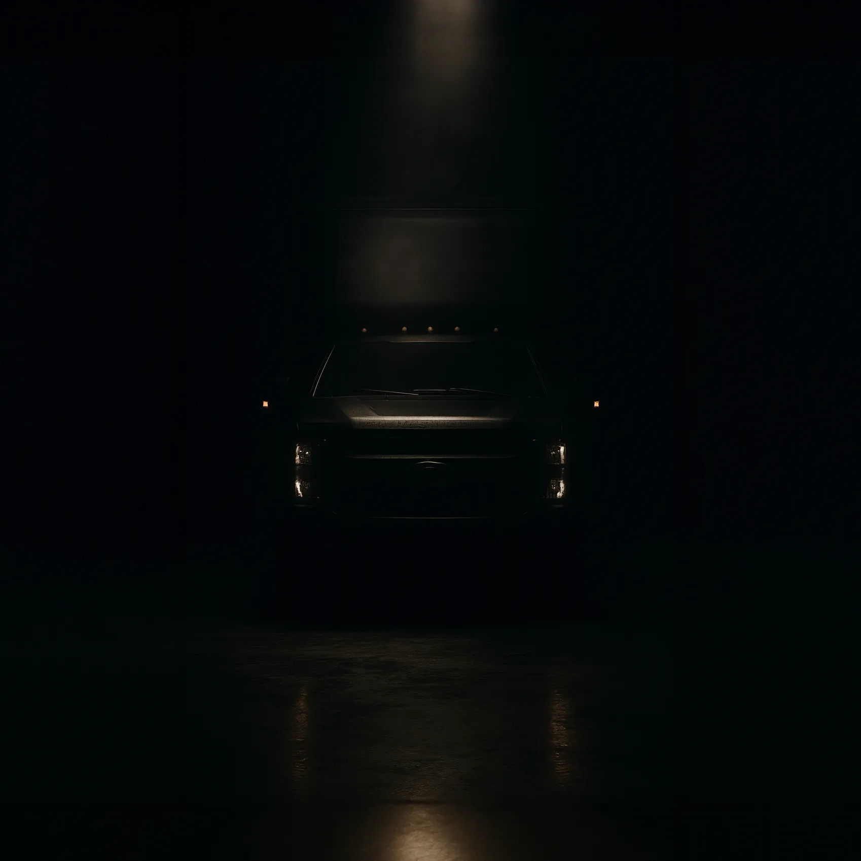 Front view of a dark truck partially illuminated by a single overhead light in a dimly lit space.